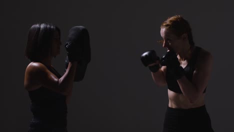 Studio-Shot-Of-Two-Mature-Women-Wearing-Gym-Fitness-Clothing-Exercising-Boxing-And-Sparring-Together-Shot-In-Profile